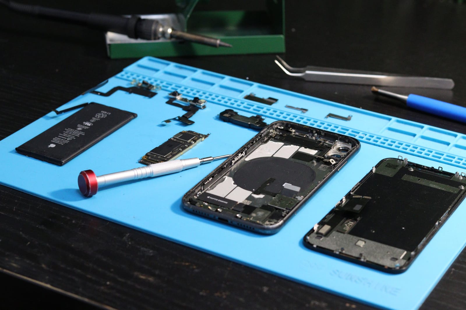 An iPhone taken apart on a repair mat with the battery, screen and logic board laid out beside repair tools